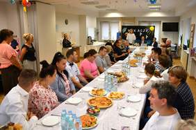 Pokaż zdjęcie: Uczestnicy biorący udział w zakończeniu roku szkolnego siedzą przy długim stole nakrytym białym obrusem. 