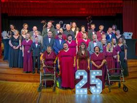 Pokaż zdjęcie: Grupa kilkudziesięciu elegancko ubranych osób ustawiona na scenie na tle czerwonych kotar; kobiety w bordowych i czarnych sukniach, mężczyźni w garniturach. Z przodu dwie podświetlane cyfry „25” oraz dwa balkoniki. Atmosfera jubileuszu lub gali.