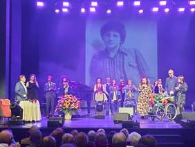 Pokaż zdjęcie: Na scenie teatru znajdują się artyści. Jedni stoją, a inni siedzą na wózkach inwalidzkich. Ponadto widać głowy osób siedzących na widowni. W tle na tablicy multimedialnej wyświetlone jest zdjęcie Krzysztofa Cwynara. 