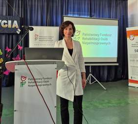 Pokaż zdjęcie: Kobieta w długim białym żakiecie, opiera rękę o mównicę z logo PFRON.