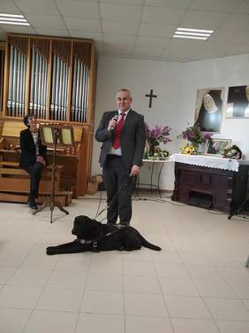 Pokaż zdjęcie: jedna osoba z psem przemawia do mikrofonu. W tle widać osobę siedzącą przy fortepianie. 