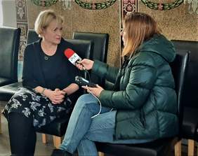 Pokaż zdjęcie: Pani Joanna Hryniewicka, z Biura Projektu Kompleksowej Rehabilitacji w Warszawie w Centrali PFRON, blondynka, udziela wywiadu dla Radia PiK, siedzi na krześle - ubrana w czarny żakiet i koszulkę oraz czarno-białą spódnicę w kwiaty,  po jej lewej stronie siedzi Pani redaktor, widoczna w połowie, która trzyma czerwony mikrofon z logo radia