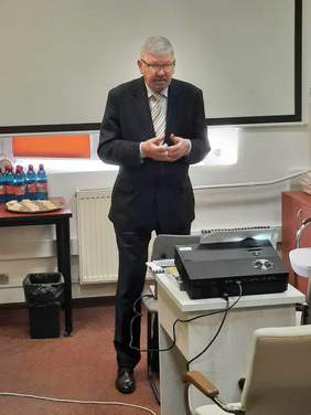 Pokaż zdjęcie: Stoi pan ubrany w garnitur i przemawia. Za nim widać tablicę multimedialną. A przed mężczyzną leży na stole rzutnik do wyświetlania prezentacji. 