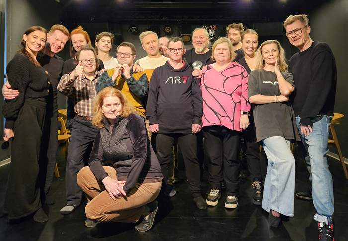 Pokaż zdjęcie: Grupowe zdjęcie zespołu zaangażowanego w spektakl „I Love the World”. Widać kilkanaście osób uśmiechających się do obiektywu w teatralnym otoczeniu. W tle scena, a na pierwszym planie różnorodne postaci – aktorzy i twórcy projektu.