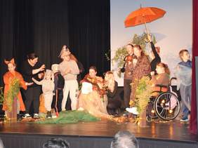 Scena teatralna podczas występu. Grupa osób w różnorodnych, kolorowych kostiumach, w tym postaci z bajek (jak lis i król), stoi i siedzi. W tle widoczny jest rekwizyt w postaci pomarańczowego parasola. Osoba na wózku inwalidzkim siedzi po prawej stronie. Na scenie leżą jesienne dekoracje.