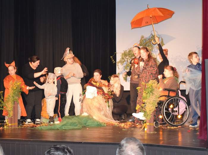 Scena teatralna podczas występu. Grupa osób w różnorodnych, kolorowych kostiumach, w tym postaci z bajek (jak lis i król), stoi i siedzi. W tle widoczny jest rekwizyt w postaci pomarańczowego parasola. Osoba na wózku inwalidzkim siedzi po prawej stronie. Na scenie leżą jesienne dekoracje.