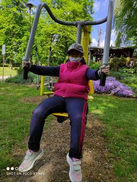 Pokaż zdjęcie: Dziewczyna w czapce z daszkiem i maseczce  ubrana w różowy bezrękawnik i dresy siedzi na drążku do ćwiczeń. W tle kwitnący ogród