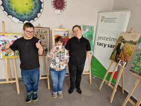 Pokaż zdjęcie: Trzy osoby z zespołem Downa pracujące w kawiarni stoją uśmiechnięte. Za nimi jest ekspozycja prac plastycznych na drewnianych sztalugach.