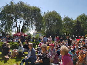 Pokaż zdjęcie: W środku parku, na trawie rozstawione są w rzędach ławki na których siedzą ludzie. W tle duże drzewa. Jest bezchmurne błękitne niebo.