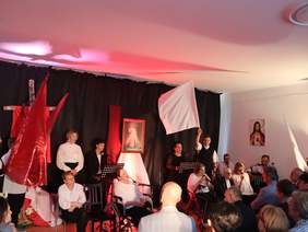 Pokaż zdjęcie: Na scenie grupa artystów, w pierwszym rzędzie sześć osób siedzi na wózkach, obok siedzi mężczyzna z akordeonem, z tyłu stoi 5 osób, śpiewają. Po lewej stronie jest drewniany krzyż. Na środku obraz Jezusa Miłosiernego.