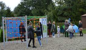 W tle widać wysokie drzewa. Na pierwszym planie plac zabaw, na którym bawią się dzieci w towarzystwie mam.