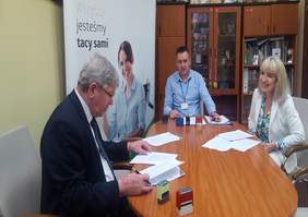 Pokaż zdjęcie: pomieszczenie w siedzibie Oddziału. Duży stól przy którym siedzą trzy osoby dwóch mężczyzn i kobieta podpisują dokumenty. W tle stoi baner z  logo PFRON