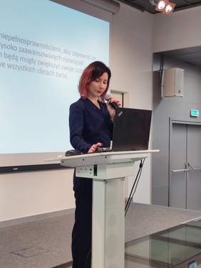 Pokaż zdjęcie: Przy białej mównicy stoi wysoka szczupła kobieta ubrana na ciemno, spogląda na laptopa.