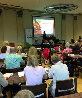 Pokaż zdjęcie: Grupa kilkunastu uczestników konferencji przysłuchuje się wystąpieniu jednego z prelegentów. Obok dużego ekranu, na którym wyświetlona jest prezentacja, stoi baner PFRON.