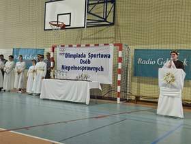 Olimpiada Sportowa Osób Niepełnosprawnych w hali sportowej. Przy bramce i stole z pucharami stoi grupa osób ubranych w białe stroje stylizowane na antyczne. Po prawej osoba przemawia przy mównicy ozdobionej wieńcem laurowym.