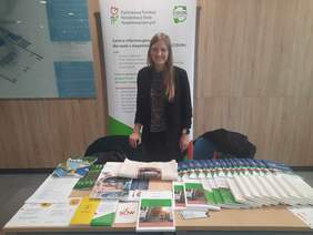 Pokaż zdjęcie: Młoda, uśmiechnięta kobieta stoi wsparta o krzesło. Przed nią ustawiony jest stół z materiałami promocyjnymi i informatorami PFRON. Za nią widoczny jest roller z tekstem opatrzonym logo PFRON i CIDON. Obok duża grafika z planem budynku.