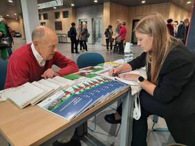 Pokaż zdjęcie: Przy stoliku siedzą dwie osoby: starszy pan i młoda kobieta. Pani opowiada o dofinansowaniach z PFRON i zapisuje infromacje dla pana na kartce.  W tle widać innych seniorów. 