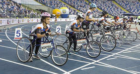 Pokaż zdjęcie: Siedem zawodniczek stoi na bieżni na linii startowej. Czekaja na sygnał do wyścigu. W tle trybuny z kibicami.