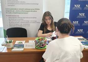 Pokaż zdjęcie: Przy biurku na którym leżą ulotki rozmawiają dwie kobiety. 