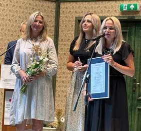 Pokaż zdjęcie: Trzy kobiety stoją na scenie podczas uroczystości. Jedna z nich trzyma dyplom i mówi do mikrofonu, druga trzyma statuetkę, a trzecia bukiet kwiatów. To przedstawicielki Stowarzyszenia My dla Innych, które otrzymało tytuł SUPER LODOŁAMACZ 2025.