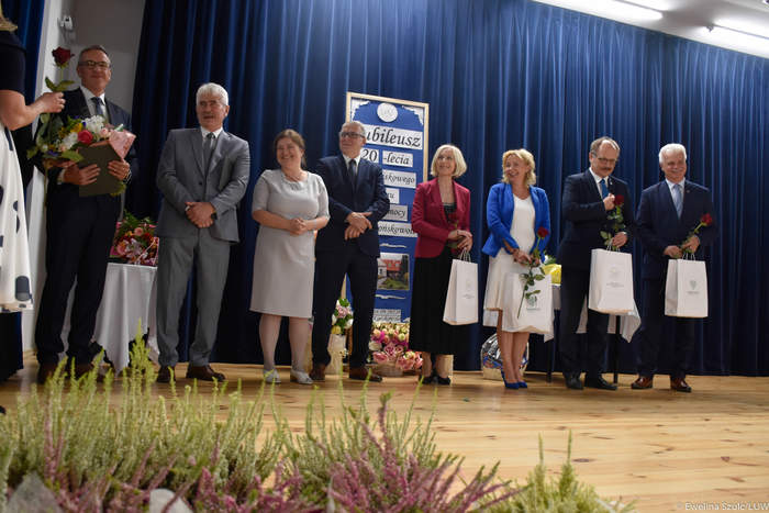 Pokaż zdjęcie: Na scenie w szeregu stoi grupa elegancko ubranych osób przyjmujących życzenia, gratulacje i pojedyncze czerwone róże