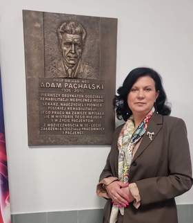 Elegancko ubrana kobieta pozuje do zdjęcia na tle tablicy pamiątkowej