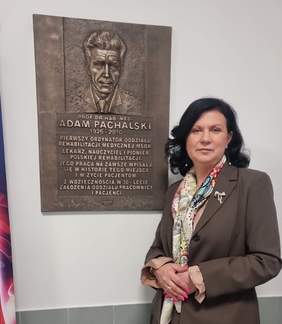 Pokaż zdjęcie: Elegancko ubrana kobieta pozuje do zdjęcia na tle tablicy pamiątkowej