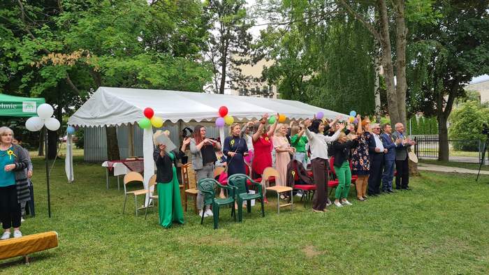 Pokaż zdjęcie: Zdjęcie w plenerze, przed namiotem ustawiono krzesełka, osoby siedzące na krzesełkach wstały i machają rękami. Część zabawy integracyjnej.