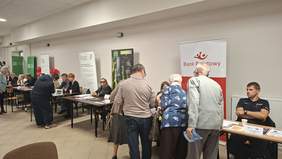 Grupa osób, kobiety i mężczyźni w różnym wieku, część stoi a część siedzi przy stolikach, w tle banery instytucji