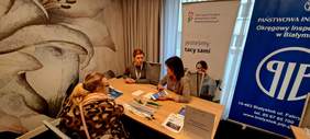 Pokaż zdjęcie: Dwie kobiety siedzą przy stole na którym leżą informatory i materiały promocyjne udzielają porady kobiecie siedzącej naprzeciwko, kobieta ubrana jest w futro w panterkę. Za kobietami z tyłu ustawione są dwa banery z logo PFRON i PIP.