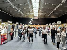 Pokaż zdjęcie: Na dużej hali znajduje się wiele ministoisk rozmieszczonych w rzędach. Pomiędzy nimi spacerują osoby.