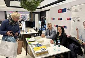 Pokaż zdjęcie: Za biurkiem, na którym lezą materiały promocyjne i informatory siedzi mężczyzna w jasnej marynarce i kobieta w ciemnej marynarce. Za nimi baner z logo PFRON i napisem wszyscy jesteśmy tacy sami. Przed nimi kobieta w zimowej kurtce patrząca na mater