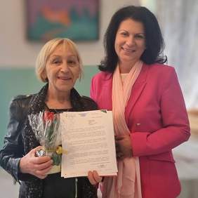 Pokaż zdjęcie: Dwie uśmiechnięte kobiety. Jedna przekazuje drugiej statuetkę i pismo gratulacyjne.