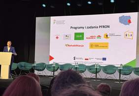 Pokaż zdjęcie: Zdjęcie z tyłu sali w kierunku ekranu, na którym jest slajd z prezentacji z napisem programy i zadania PFRON.