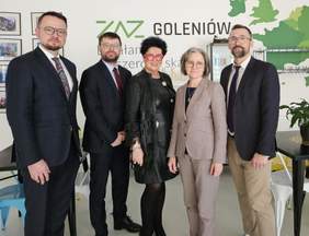 Pokaż zdjęcie: Na zdjęciu jest 5 osób, w środku dwie Panie. Pozują do zdjęcia. W tle Zielono czarny napis ZAZ Goleniów i grafika z zieloną mapą