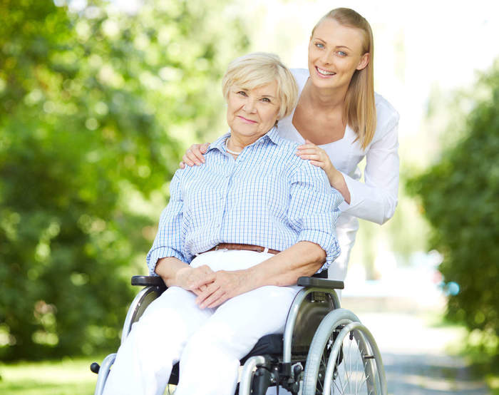 starsza osoba na wózku dla osoby niepełnosprawnej, za nią osoba z gestem opiekuńczości