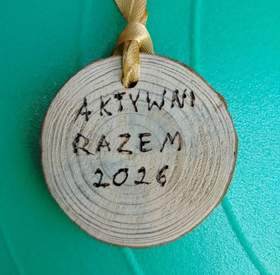 Drewniany medal w kształcie krążka z widocznymi słojami drewna na tle w intensywnym turkusowym kolorze. U góry medalu jest przewleczona żółtawa wstążka. Na środku medalu widnieje odręczny napis wypalony w drewnie: Aktywni Razem 2026.