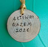 Drewniany medal w kształcie krążka z widocznymi słojami drewna na tle w intensywnym turkusowym kolorze. U góry medalu jest przewleczona żółtawa wstążka. Na środku medalu widnieje odręczny napis wypalony w drewnie: Aktywni Razem 2026.