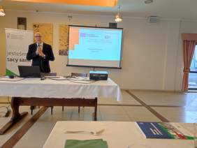 Pokaż zdjęcie: Za stołem ma którym leży laptop stoi Mężczyzna w okularach w dłoniach trzyma mikrofon. Za nim na ekranie wyświetlona jest prezentacja.