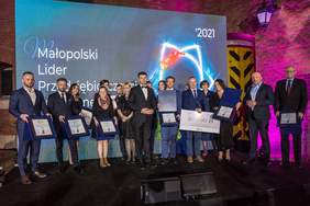 Pokaż zdjęcie: Na scenie stoją Laureaci, przedstawiciele Kapituły wręczają nagrody laureatom. Wszyscy są elegancko ubrani.