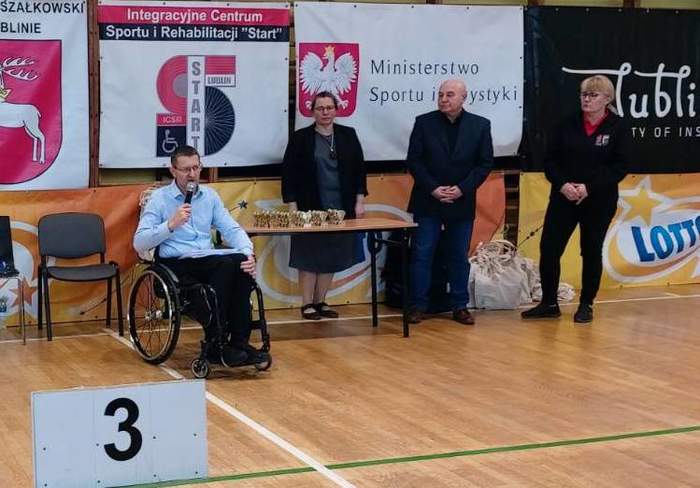 Pokaż zdjęcie: W hali sportowej wyczytywana jest lista zwycięzców, osoba na wózku inwalidzkim trzyma mikrofon, obok stoi kobieta i mężczyzna w ciemnym garniturze.