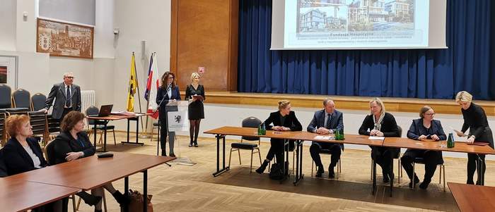 Pokaż zdjęcie: Na dużej Sali stoi długi stół. Za nimi siedzą trzy kobiety i mężczyzna w garniturze. Po lewej stronie mównice przy niej dwie elegancko ubrane kobiety. Za nimi scena z ciemną kurtyną nad sceną wisi telebim z projektem obiektu.