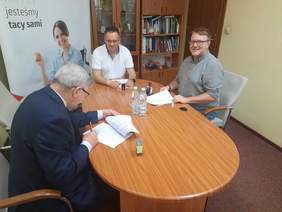 Pokaż zdjęcie: Przy owalnym stole siedzi trzech panów. Jeden z mężczyzn ubrany w garnitur podpisuje dokumenty. W tle widać regał z księżkami i odznaczeniami. 