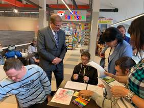 Pokaż zdjęcie: Trzech niepełnosprawnych artystów siedzi przy stoliku i rysuje na kartkach. 