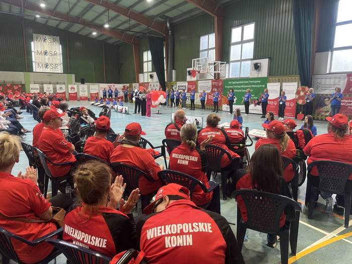 Pokaż zdjęcie: W dużej hali sportowej znajduje się grupa kilkudziesięciu osób, wśród której są sportowcy ubrani w jednolite stroje. Na obrzeżach hali ustawiona jest duża ilość banerów informujących o wydarzeniu oraz o podmiotach finansujących przedsięwzięcie.