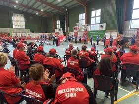 Pokaż zdjęcie: W dużej hali sportowej znajduje się grupa kilkudziesięciu osób, wśród której są sportowcy ubrani w jednolite stroje. Na obrzeżach hali ustawiona jest duża ilość banerów informujących o wydarzeniu oraz o podmiotach finansujących przedsięwzięcie.