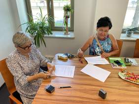 Pokaż zdjęcie: Przy drewnianym stole siedzą dwie kobiety w okularach ubrane we wzorzyste sukienki. Na stole leżą dokumenty, okulary, książka,  pieczątki i cukierki. Kobieta z lewej otwiera umowę, kobieta z prawej trzyma długopis w ręku nad umową, patrzy w obie