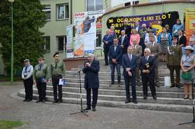 Pokaż zdjęcie: Na schodach przed budynkiem stoją ludzie. Nad nimi wisi transparent z napisem: 36 Ogólnopolski Zlot Drużyn Nieprzetartego Szlaku ZHP. Jeden mężczyzna przemawia do mikrofonu. 