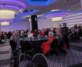 Pokaż zdjęcie: Zdjęcie przedstawia elegancką salę w Hanza Tower w Szczecinie podczas spotkania wigilijnego Stowarzyszenia Amazonek „AGATA”. Sala wypełniona jest kobietami siedzącymi przy okrągłych stołach, ozdobionych zielonymi serwetkami i świątecznymi dekoracjami. Na suficie widoczne są nowoczesne, podświetlane żyrandole w odcieniach fioletu, co tworzy przyjemny, ciepły nastrój. W tle widać choinkę i osoby zaangażowane w organizację wydarzenia. Część uczestniczek robi zdjęcia lub słucha występów w skupieniu, co podkreśla wyjątkowy charakter tej uroczystości.