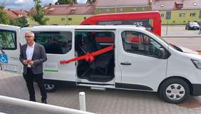 Pokaż zdjęcie: Kierownik WTZ Marian Wilkowski na tle nowego samochodu z przewiązaną czerwoną wstążką z kokardą na drzwiach.  Po boku samochodu są naklejki z logiem Państwowego Funduszu Rehabilitacji Osób Niepełnosprawnych.